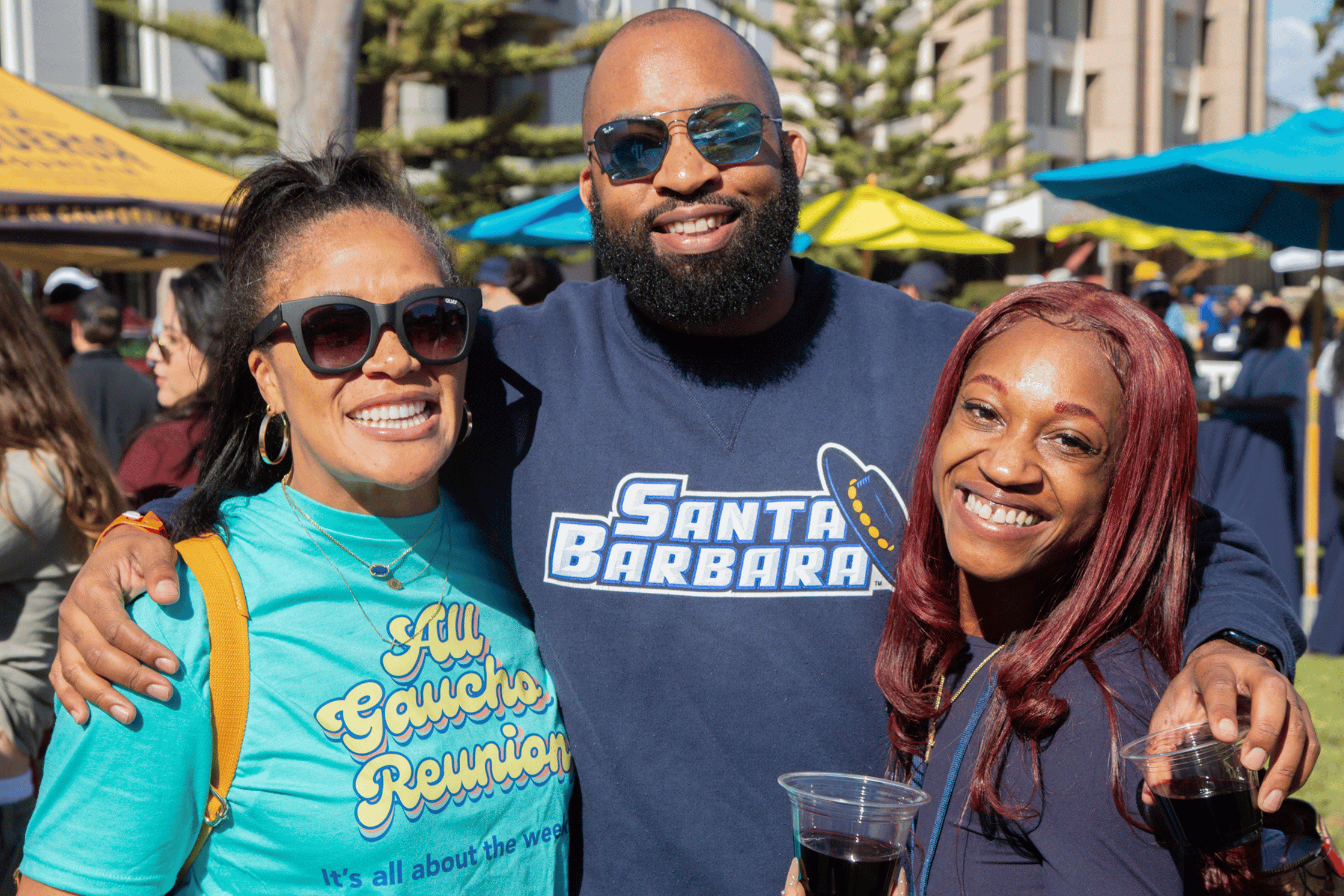 Three smiling alums embrace warmly while holding drinks. 