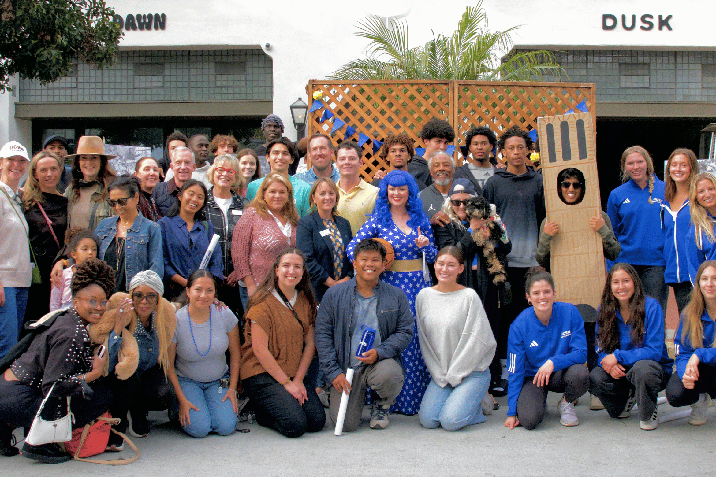A large group of people poses together outdoors alongside festive characters, including someone in a cardboard tower costume.
