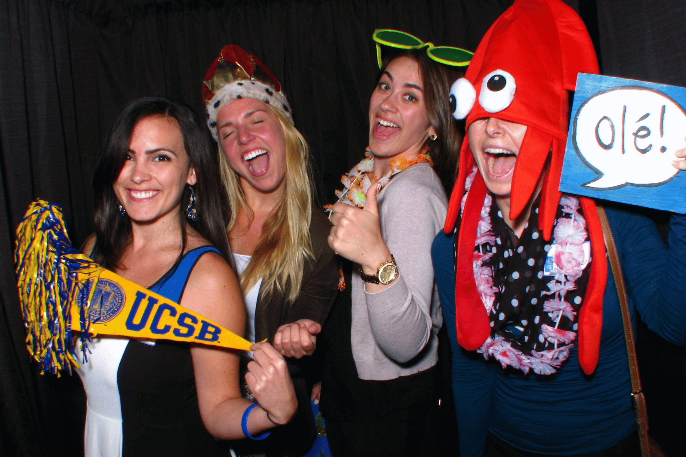 Four alums playfully pose in a photo booth with fun props, including a UCSB pennant, a squid hat, and an "Olé!" sign.