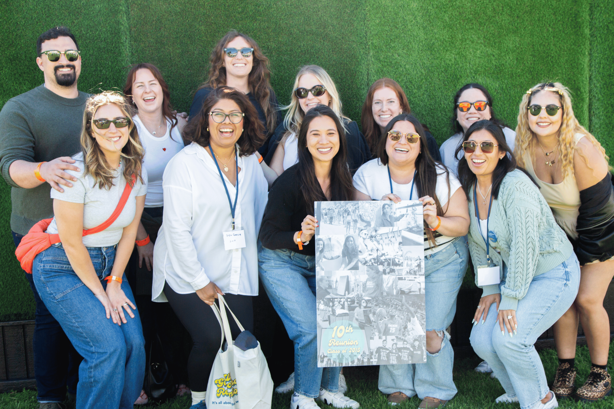 A happy group of alumni smiles together in front of a green hedge wall while holding up a "10th Reunion Class of 2014" photo poster.