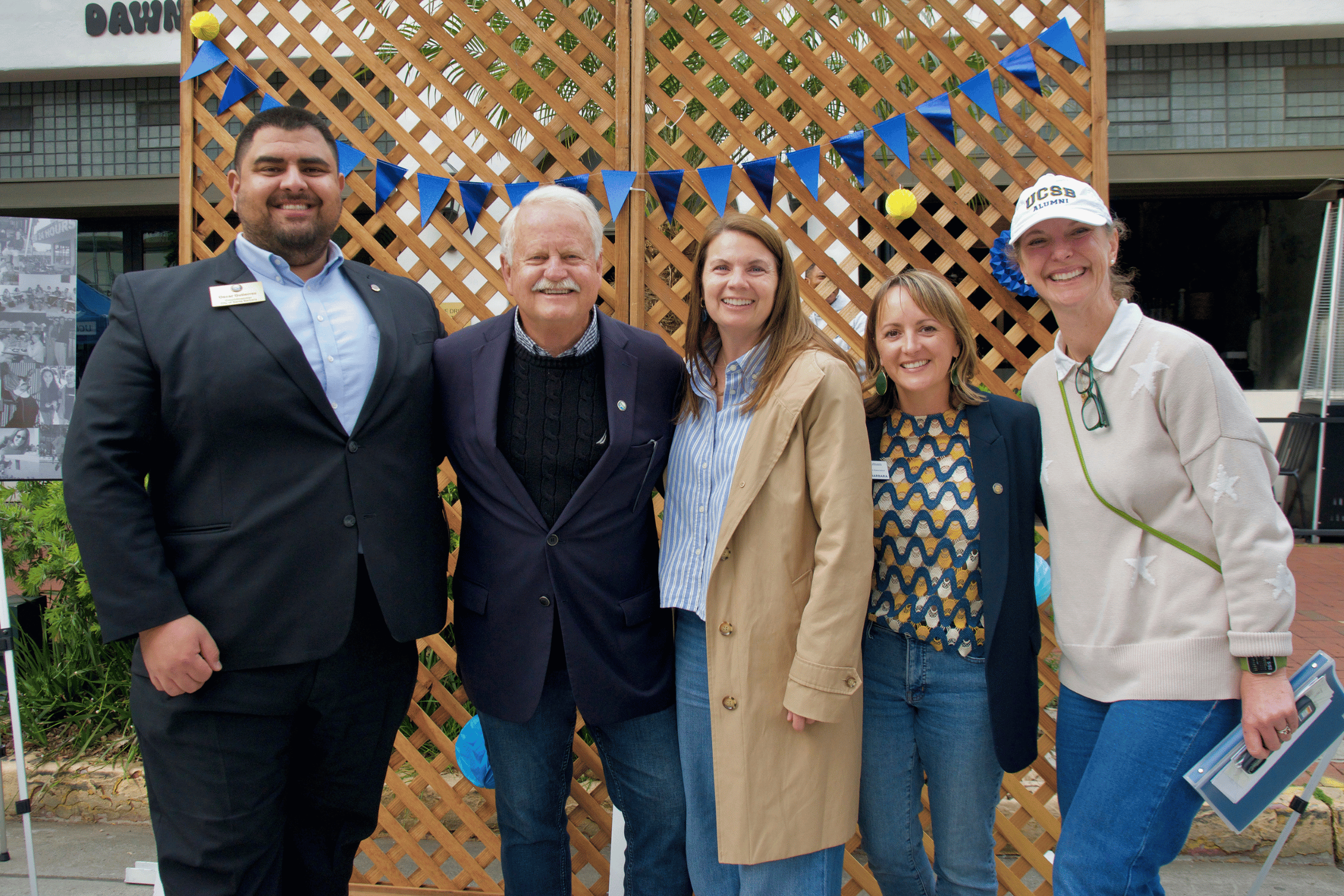 Five people stand side-by-side and smile for a photo in front of a wooden lattice backdrop decorated with blue festive banners.
